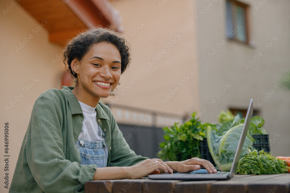 Smiling woman vegetables farm owner working on laptop with orders on background of building